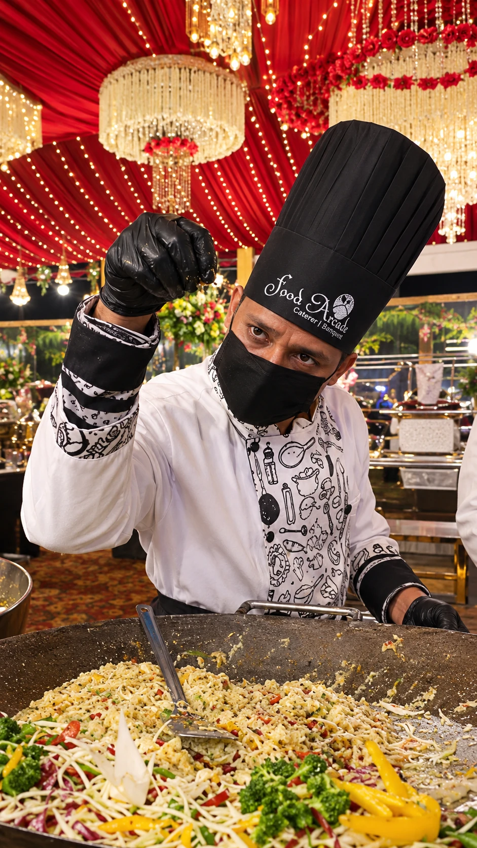 Chef in a black hat cooking noodles and vegetables in a large wok beneath red drapes and chandeliers