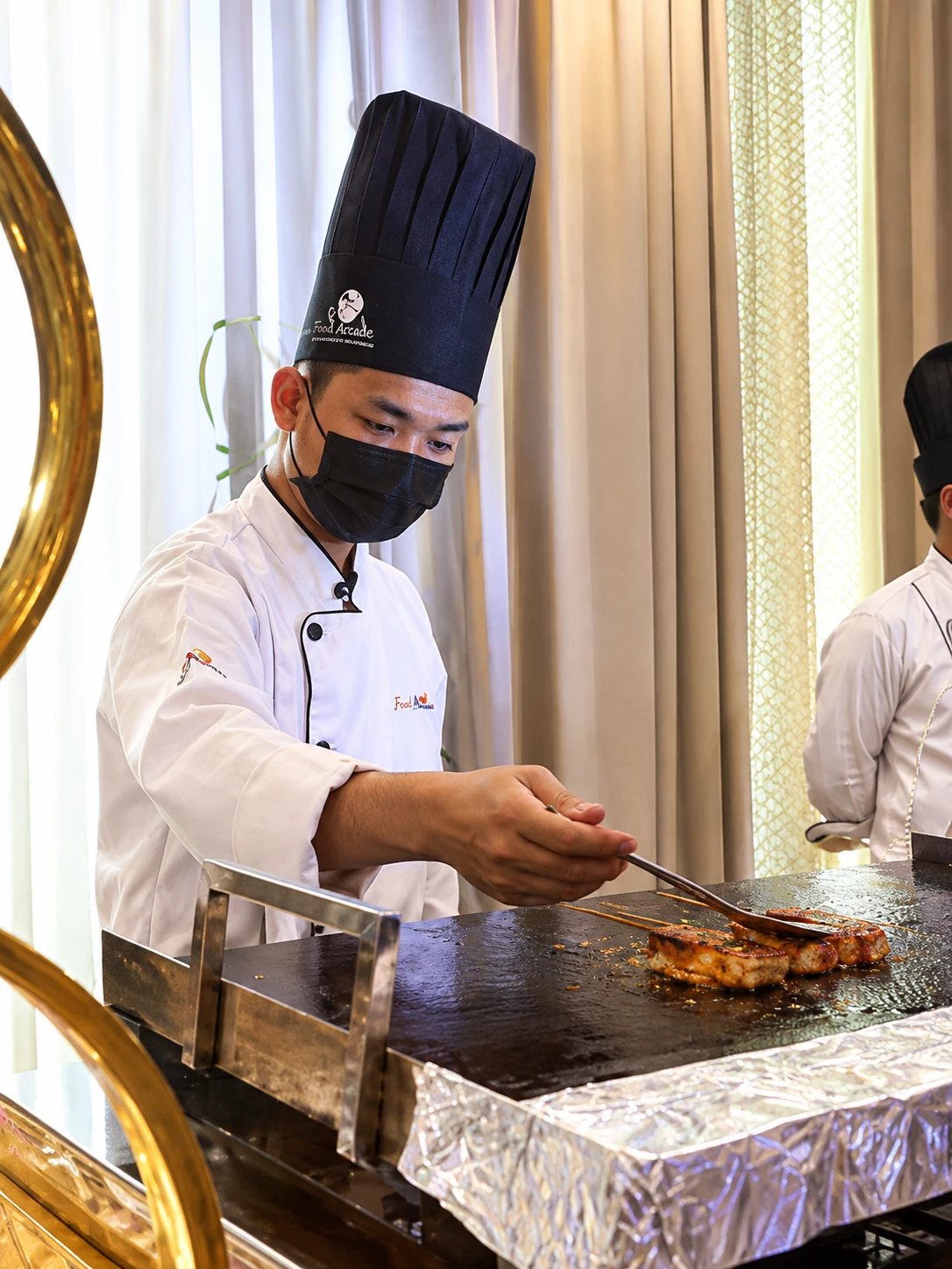 Chef in a tall hat grilling marinated pieces on a flat griddle at a live counter