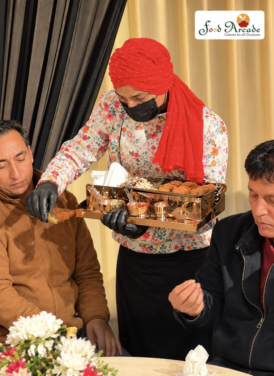 Server in a red turban and floral jacket presenting starters from a gold tray to seated guests
